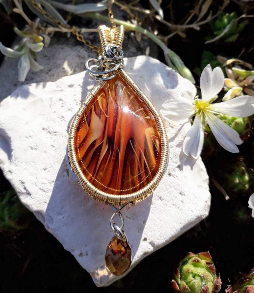 pendentif verre murano de couleur ambre tissé au fil acier inoxydable et au laiton avec une goutte de swarovski photo extérieure prise sur une pierre blanche accompagnée d'une fleur